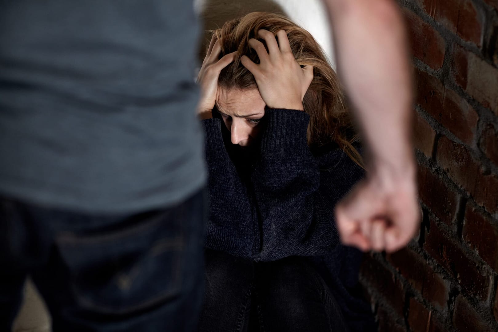 A distressed individual sitting against a wall with their head in their hands, while a clenched fist looms in the foreground, indicating fear and possible threat.