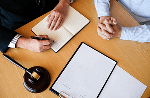 Two people sitting at a table discussing legal matters, one writing notes in a notebook while the other clasps their hands, with a gavel and legal documents in view. Finding the Right Family Law Lawyers Toronto for Your Legal Needs.